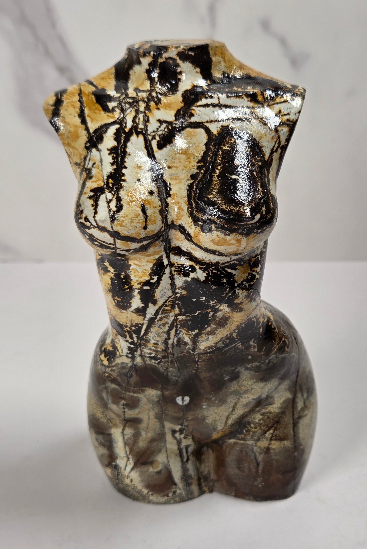 A ceramic female torso sculpture with a glossy, marbled finish in black, brown, and beige, set against a light gray marble-patterned background.