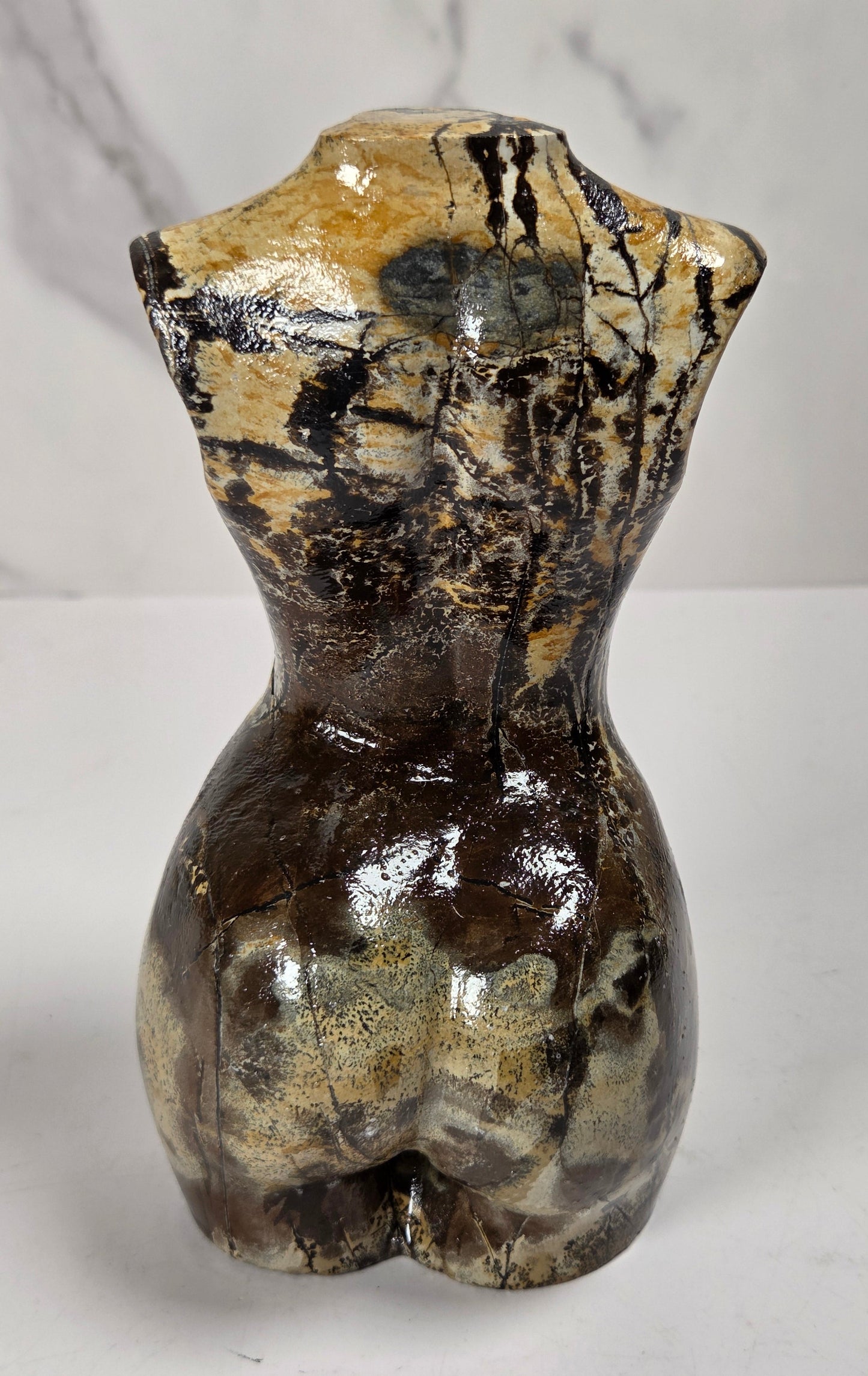 A small sculptural torso-shaped carving with a glossy marbled finish in beige, brown, and black, displayed on a white surface against a light gray marble-patterned background.