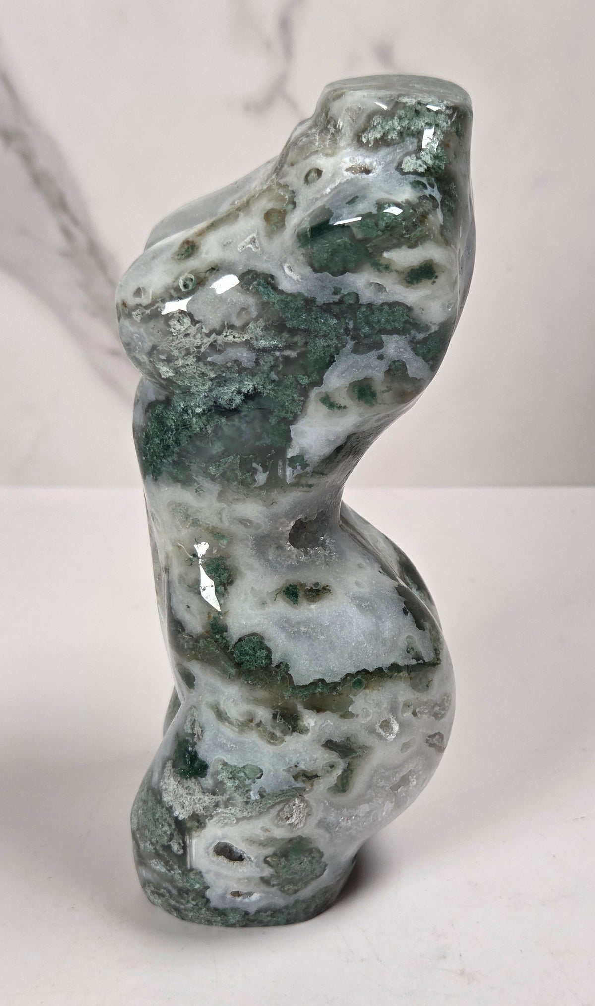 A small sculptural carving made from polished green and white marbled stone, standing upright on a white surface against a light gray marble-look background.
