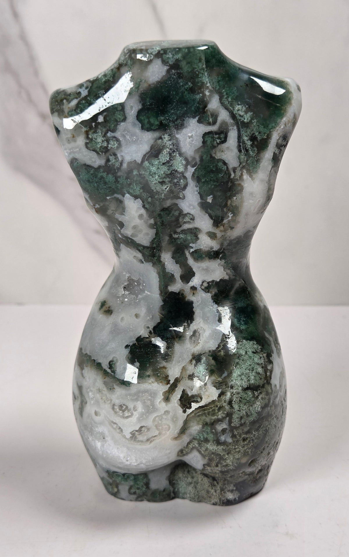 A small sculptural torso-shaped object made from a mottled green and white stone or mineral, photographed on a light surface against a pale marble-like background.