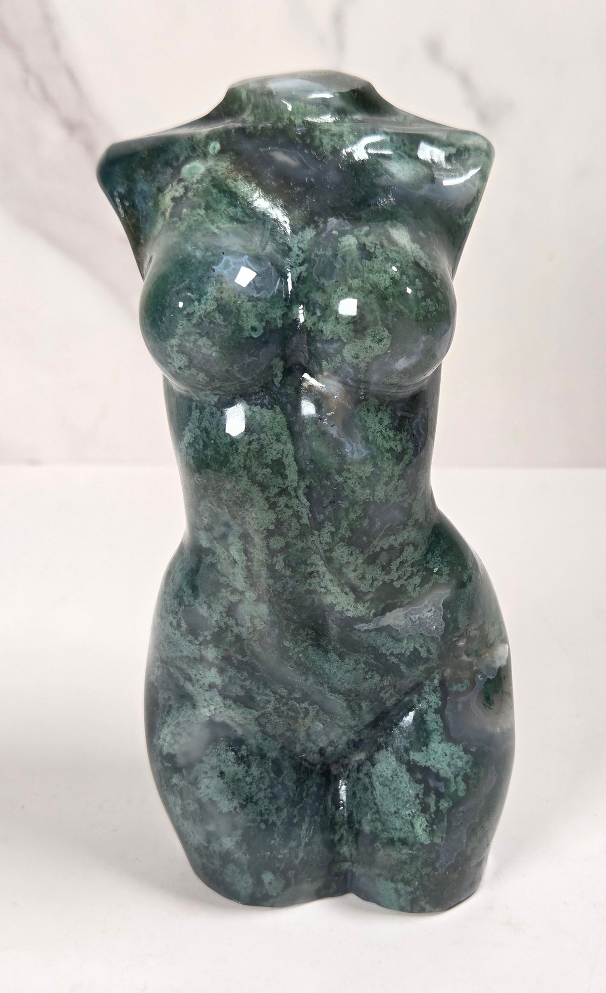 A glossy green stone torso sculpture with natural marbled texture, standing upright on a white surface against a light gray marble-patterned background.