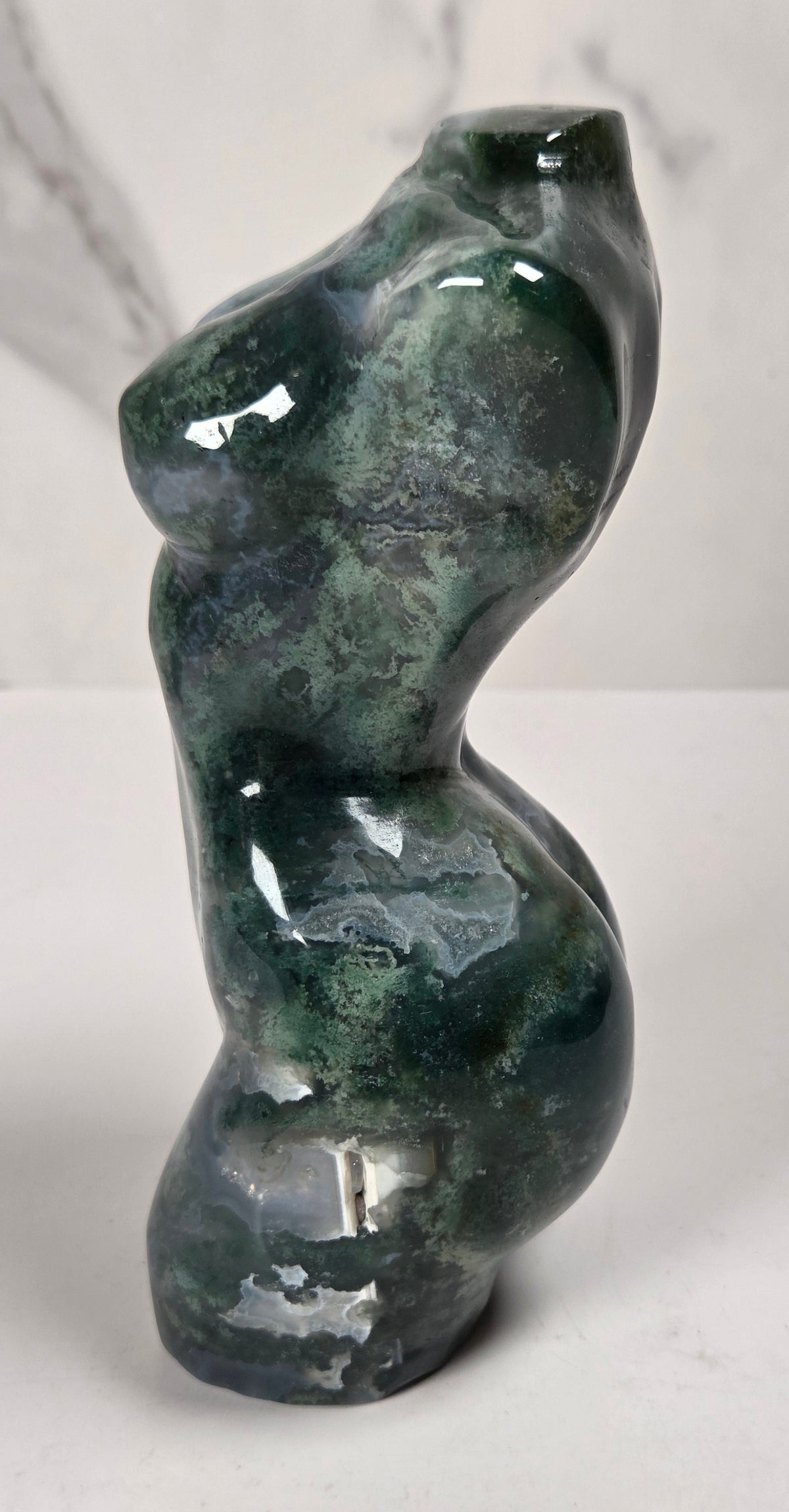 A small abstract sculpture shaped like a stylized female torso, made from glossy dark green stone with natural marbling and highlights, set against a light gray marble-patterned background.