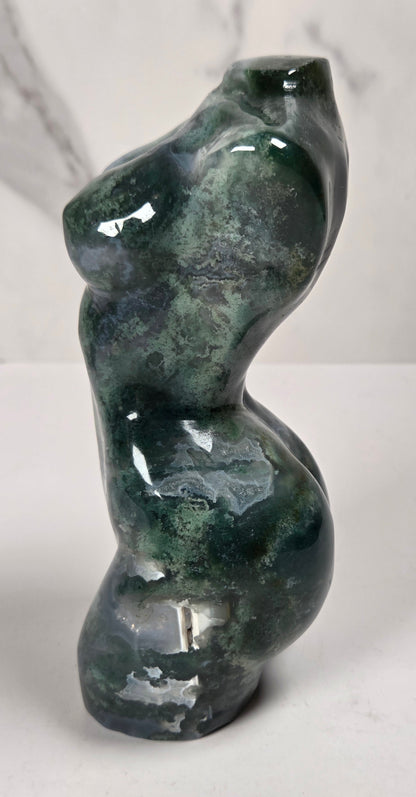 A small abstract sculpture shaped like a stylized female torso, made from glossy dark green stone with natural marbling and highlights, set against a light gray marble-patterned background.