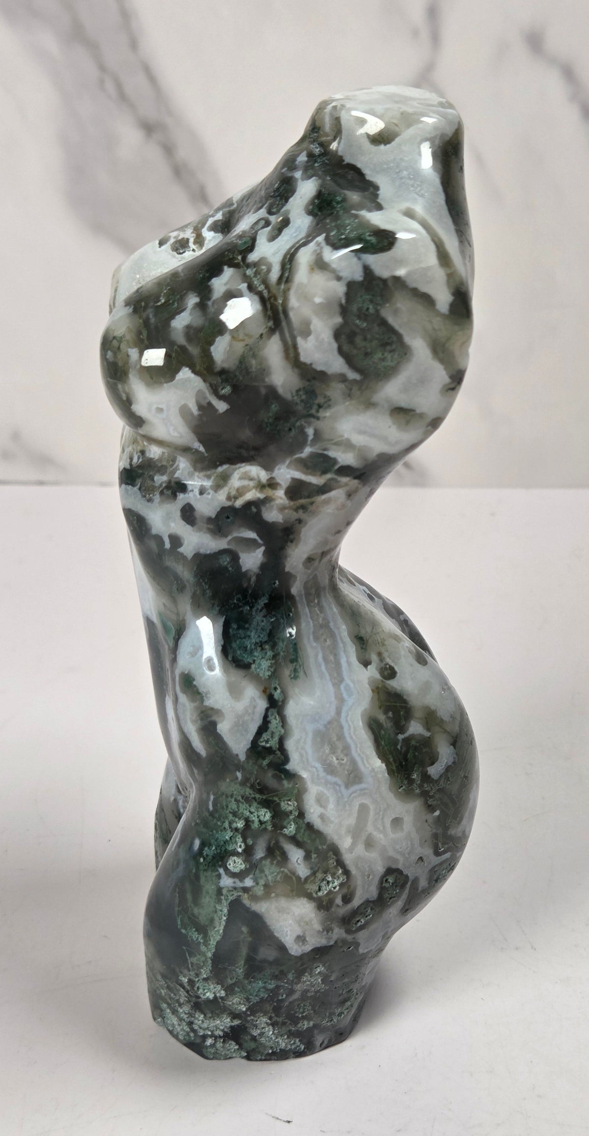 A polished natural stone sculpture shaped like a stylized female torso, featuring marbled patterns of dark green, white, and gray, set against a light gray marble-patterned background.