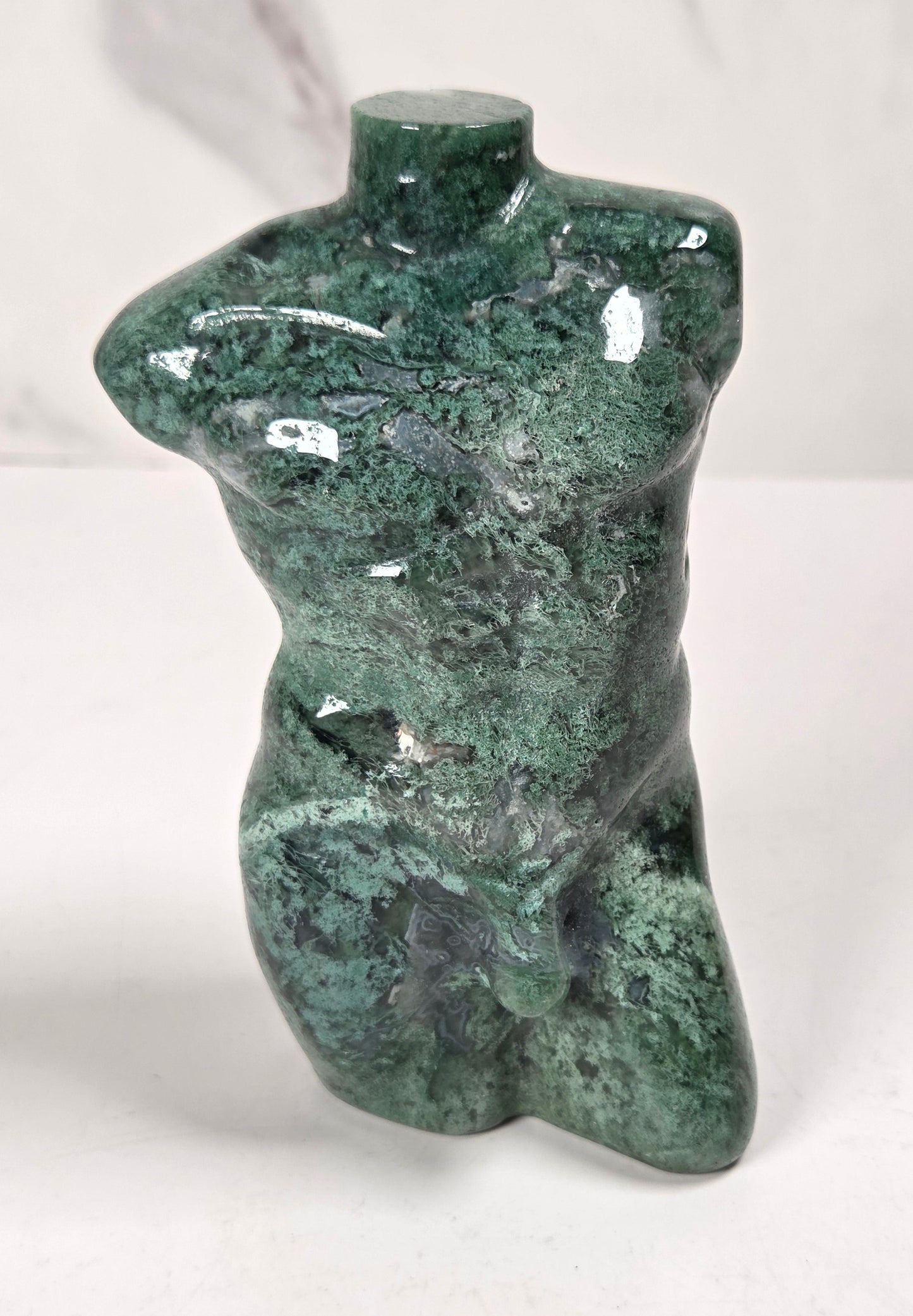 A small glossy green stone sculpture shaped like a stylized torso, photographed on a white surface against a light gray marble-look background.