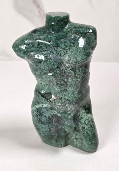 A small glossy green stone sculpture shaped like a stylized torso, photographed on a white surface against a light gray marble-look background.