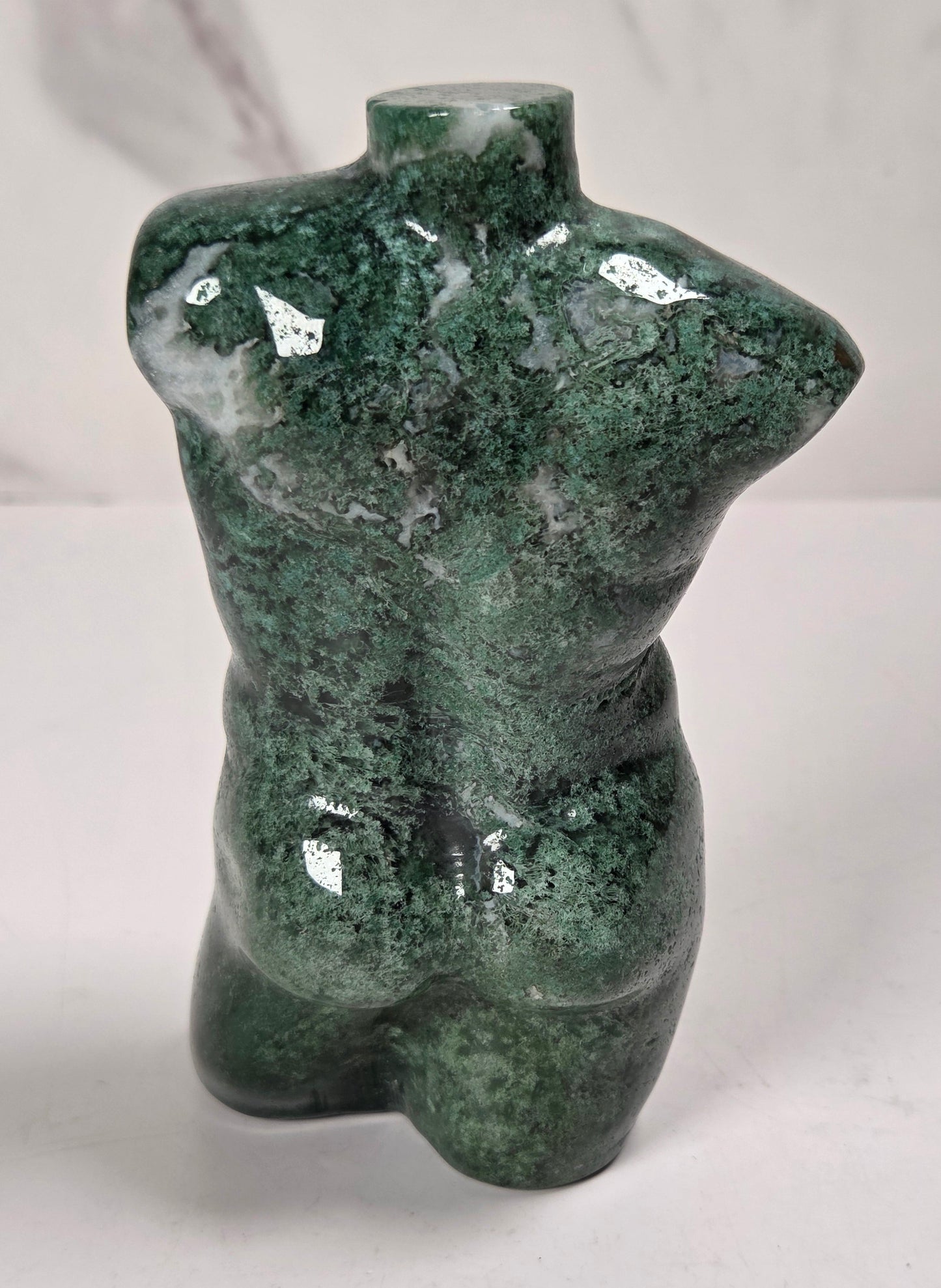 A small sculptural torso figurine made of polished green stone with natural marbling, standing on a white surface against a light gray marble-patterned background.