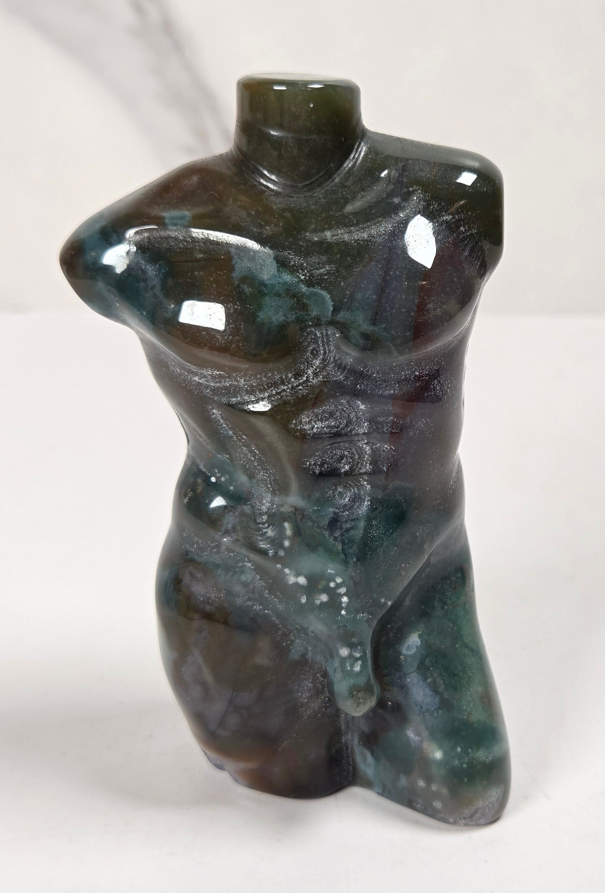 A small sculptural vase shaped like a stylized torso, made from polished dark stone with green, blue, and brown marbling, set against a light marble-patterned background.