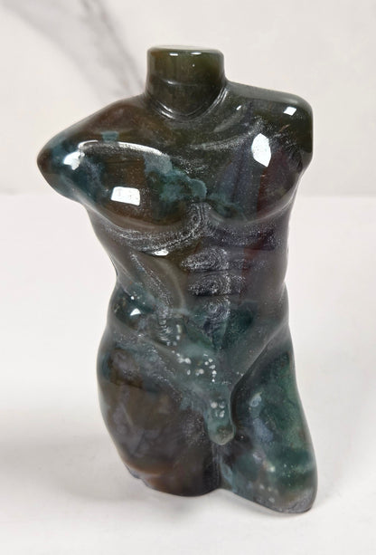 A small sculptural vase shaped like a stylized torso, made from polished dark stone with green, blue, and brown marbling, set against a light marble-patterned background.
