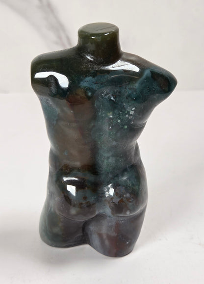 A small glossy torso-shaped decorative sculpture or figurine in dark blue-green and brown marbled tones, photographed against a light marble-patterned background.