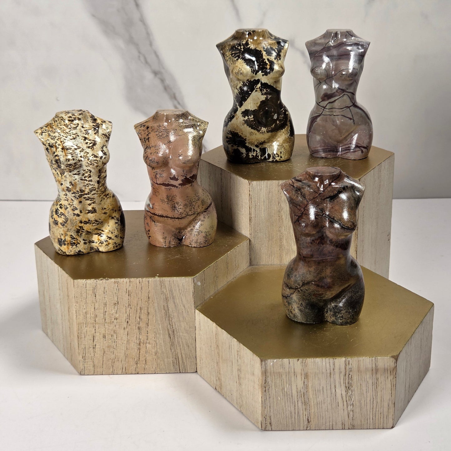 Five small torso-shaped sculptures in varied marbled and metallic finishes are displayed on geometric wooden pedestals against a light marble-patterned background.