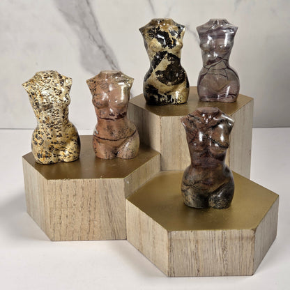 Five small torso-shaped sculptures in varied marbled and metallic finishes are displayed on geometric wooden pedestals against a light marble-patterned background.