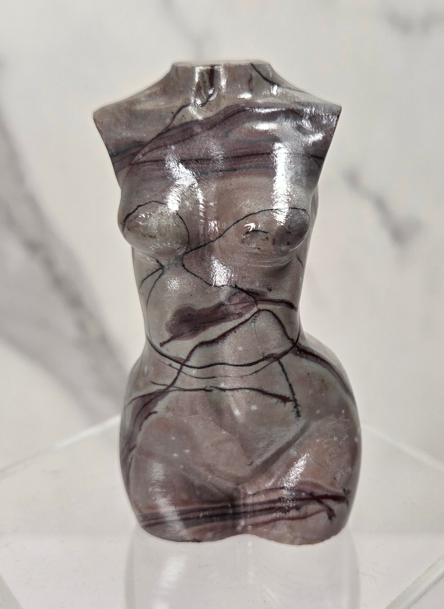 A small glossy abstract female torso sculpture in smoky translucent gray with dark veining, displayed on a clear acrylic stand against a light marble-patterned background.