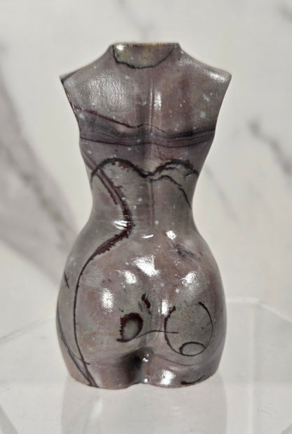 A small sculptural torso-shaped vase or candle holder made from polished stone with a glossy, metallic gray and purple marbled finish, displayed on a clear acrylic block against a light gray marble-patterned background.