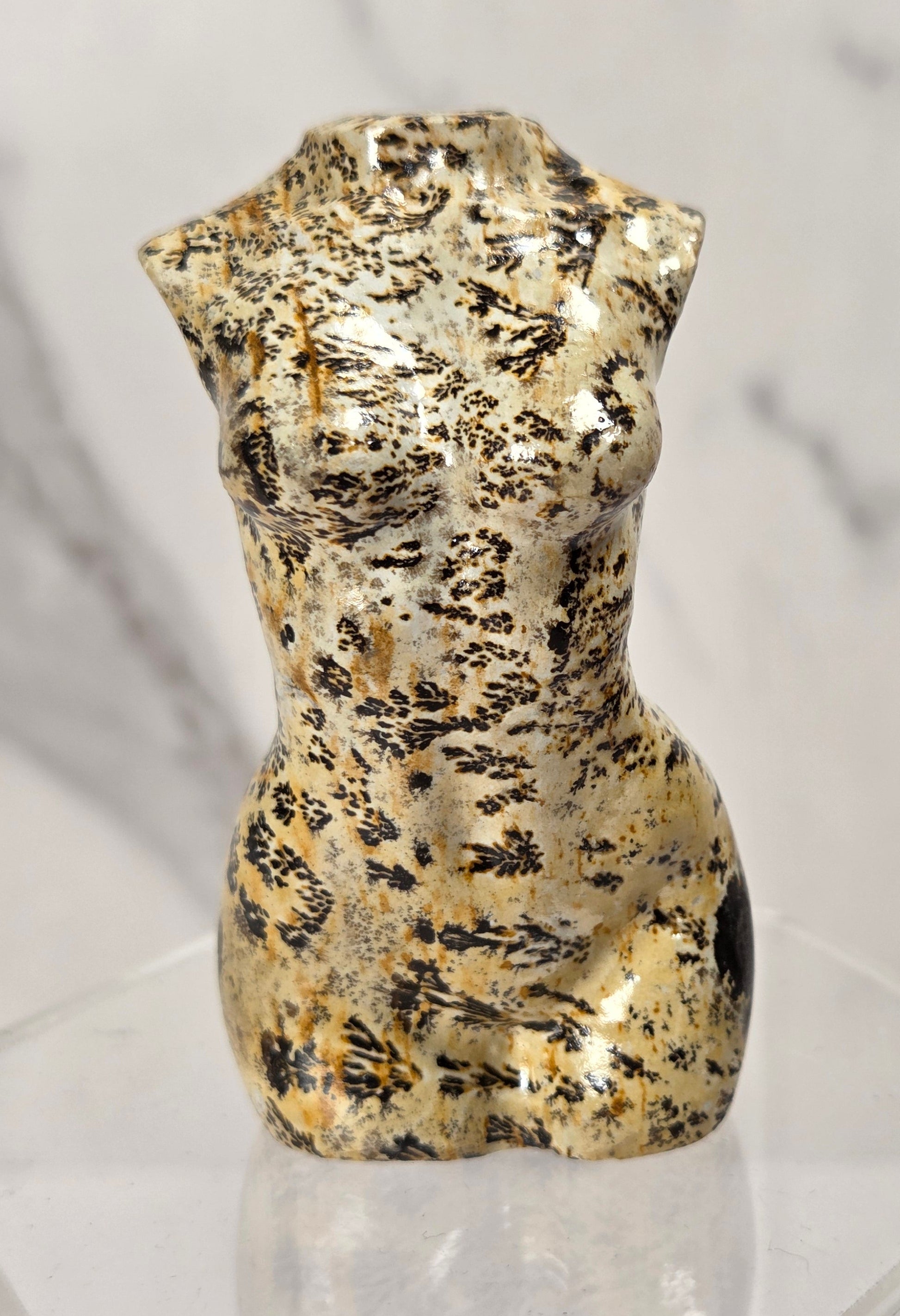A small sculptural torso figurine with a polished gold and black mottled finish, displayed on a clear acrylic pedestal against a light marble-patterned background.