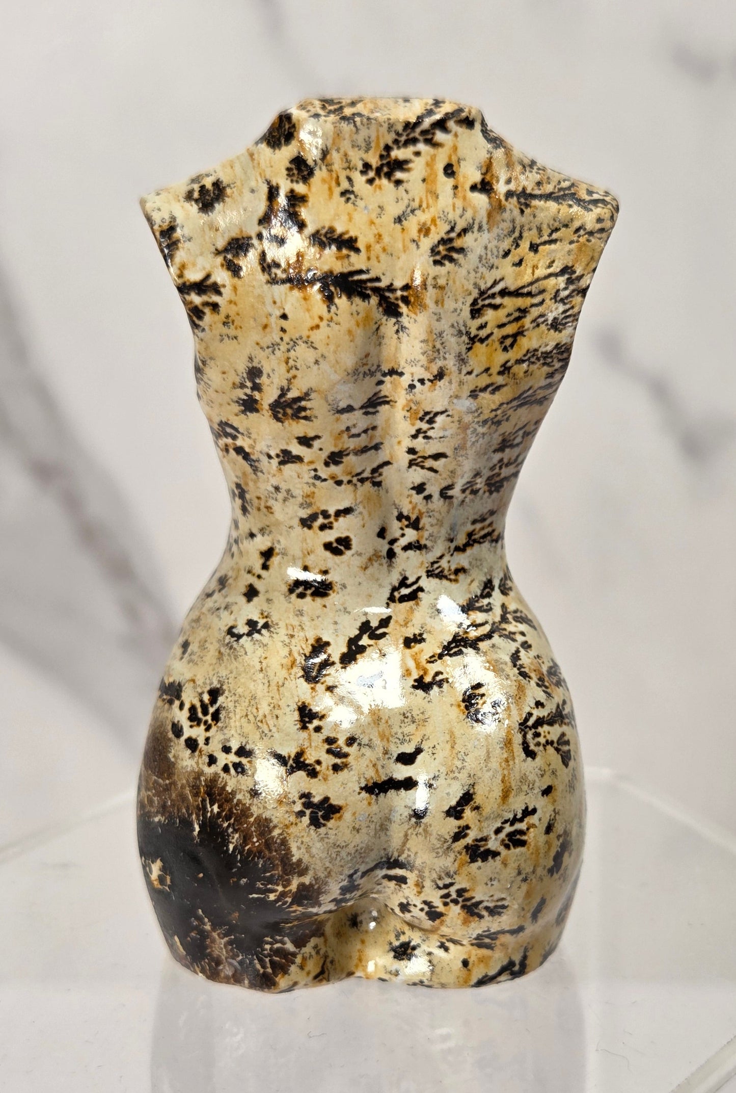 A small abstract sculptural torso or bottle-like form with a metallic gold and black marbled pattern, displayed on a clear acrylic pedestal against a light marble-patterned background.
