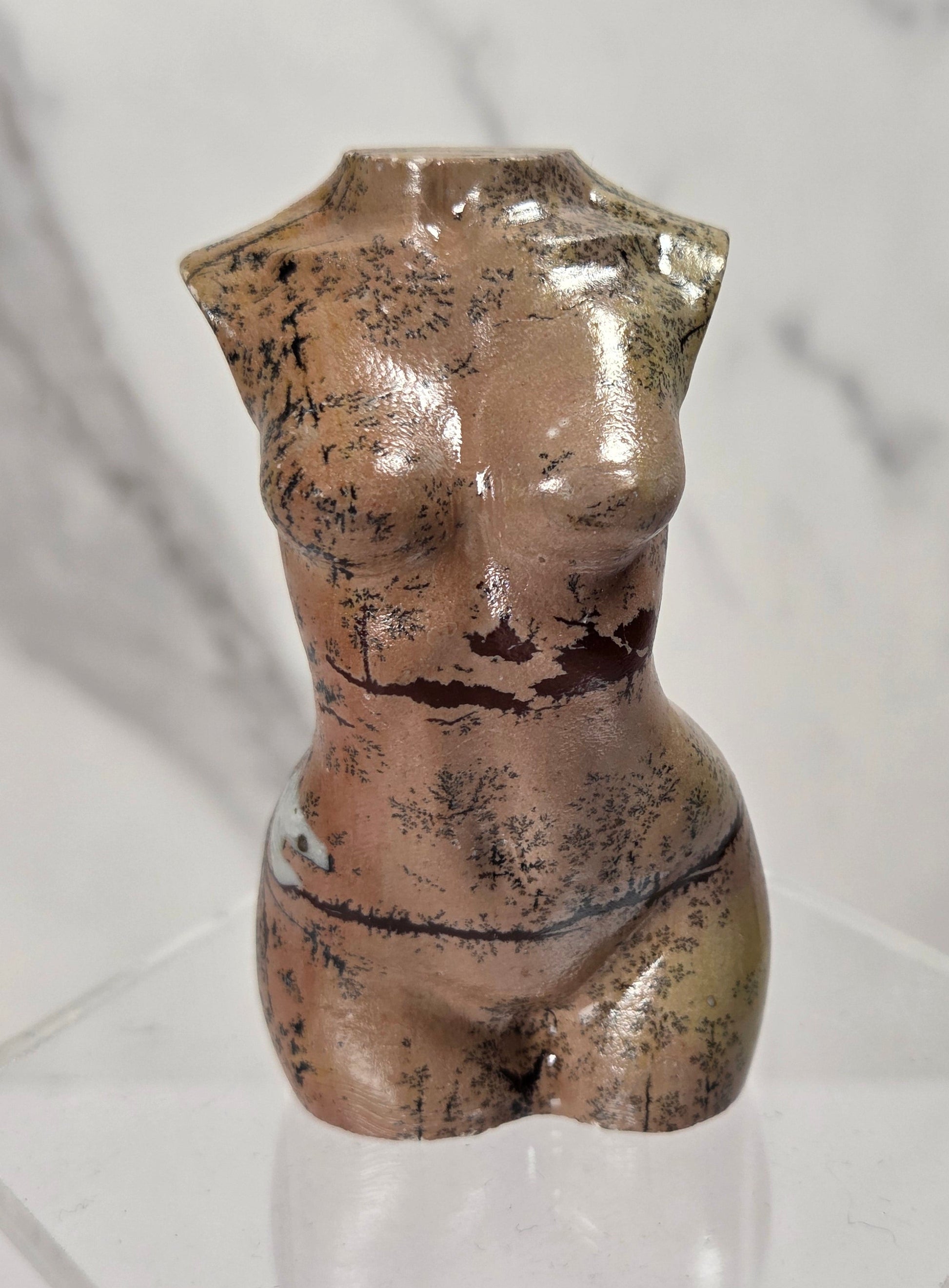 A small glossy ceramic female torso sculpture with a distressed, mottled brown and gold finish, displayed on a clear acrylic hexagonal pedestal against a light gray marble-patterned background.