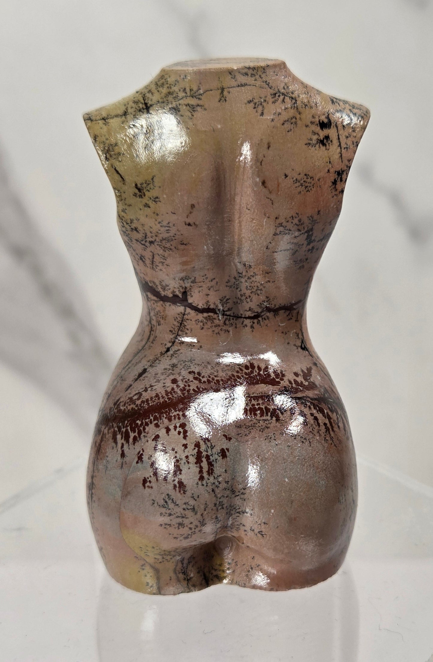 A small sculptural bust of a female torso with a glossy finish, featuring a mottled brown, bronze, and gold glaze with dark speckled details, displayed on a clear acrylic hexagonal pedestal against a light gray marble background.