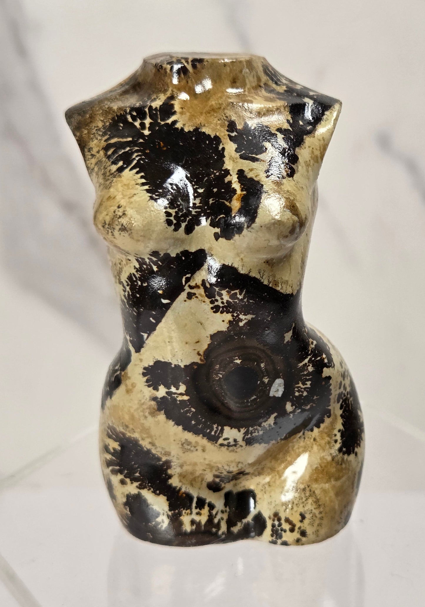 A small glossy ceramic figurine shaped like a stylized female torso, decorated with black and beige marbled patterns, displayed on a clear acrylic stand against a light marble-patterned background.