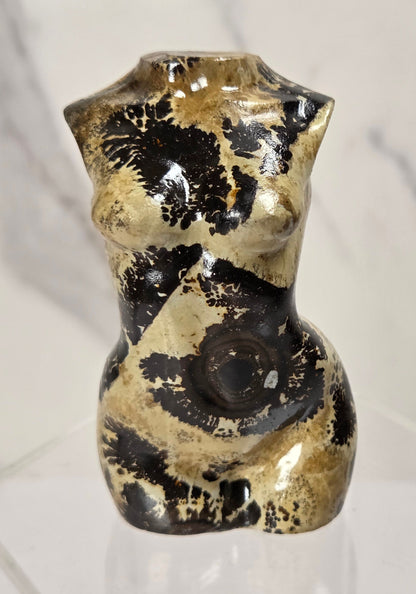 A small glossy ceramic figurine shaped like a stylized female torso, decorated with black and beige marbled patterns, displayed on a clear acrylic stand against a light marble-patterned background.