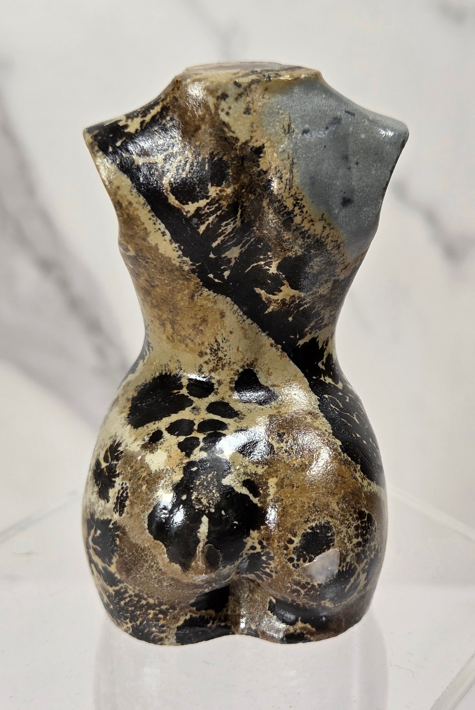 A small abstract torso sculpture with a glossy marbled finish in black, tan, and gray, displayed on a clear acrylic stand against a light marble-patterned background.