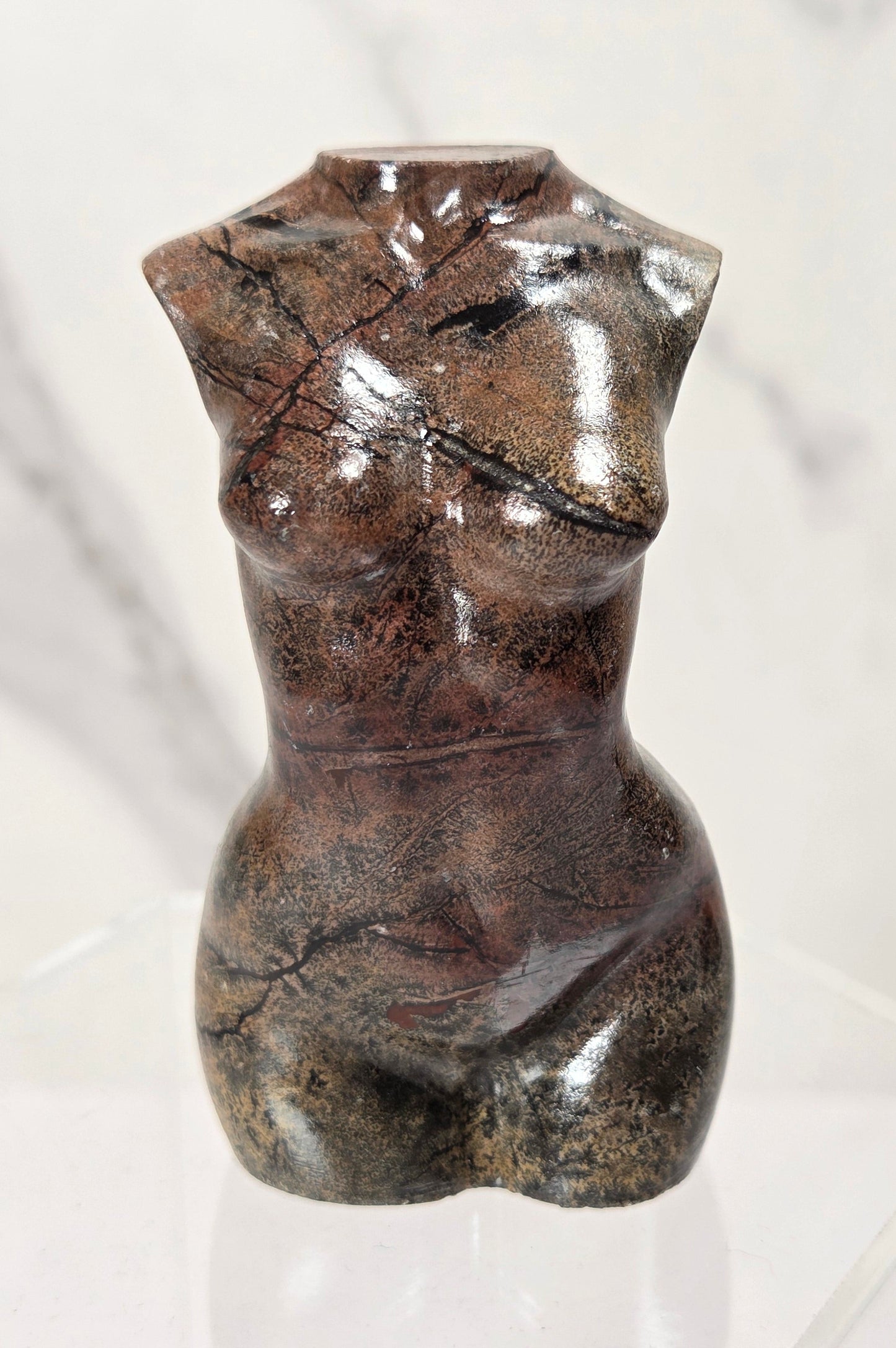 A small sculptural torso vase made from polished brown stone with natural marbling and dark fissures, displayed on a clear acrylic stand against a light marble-patterned background.