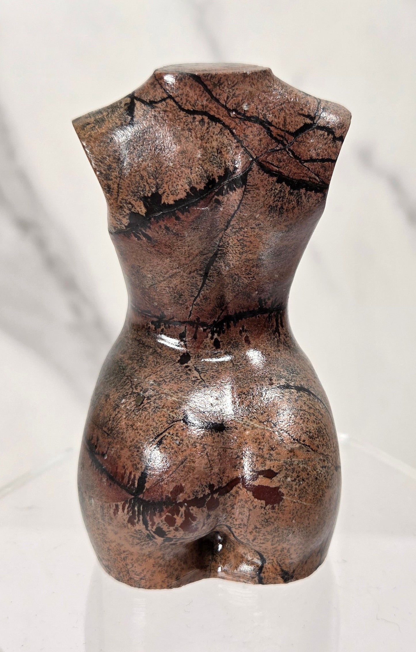 A small glossy brown sculpture or figurine with a cracked stone-like surface, displayed on a clear acrylic stand against a light marble-pattern background.