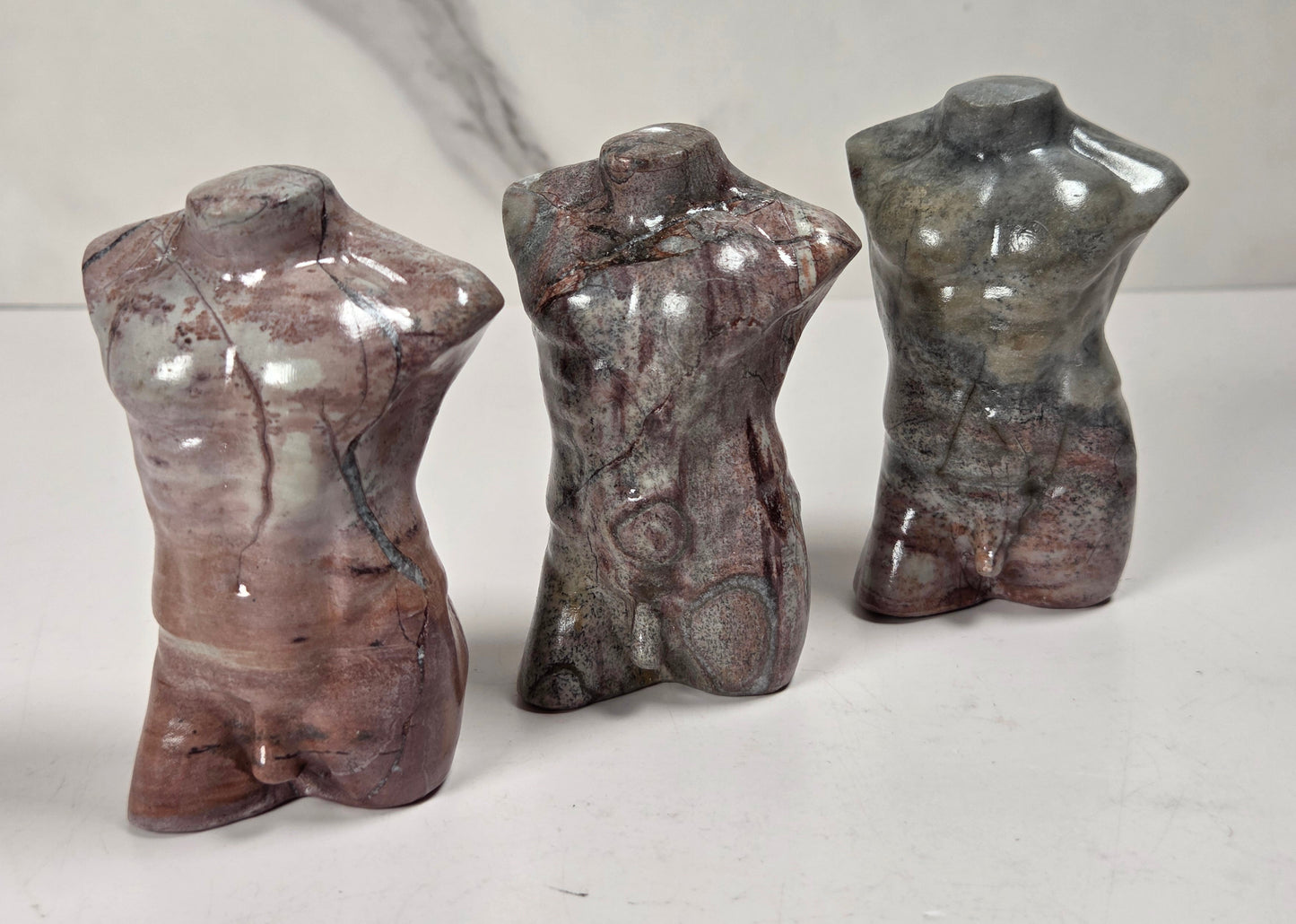 Three small polished stone torso sculptures standing upright on a white surface against a light gray background.