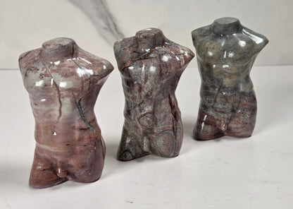 Three small polished stone torso sculptures standing upright on a white surface against a light gray background.