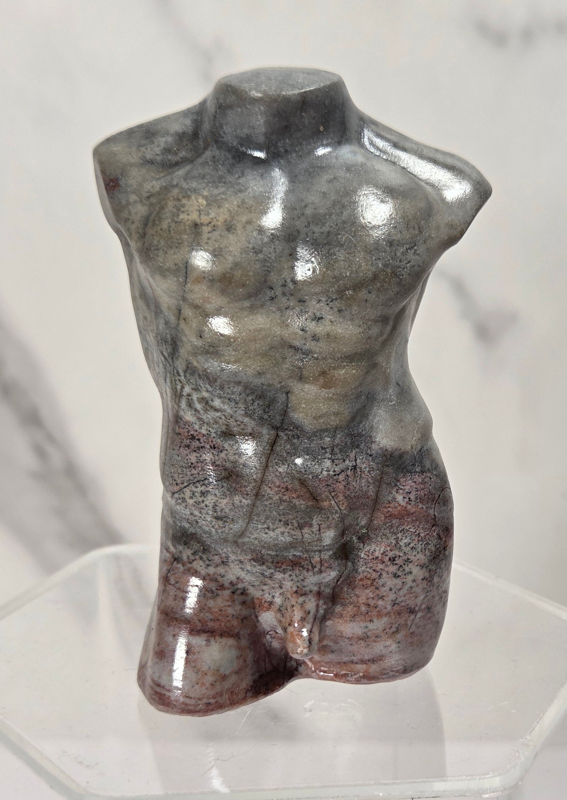 A polished stone torso sculpture with a mottled gray, brown, and reddish surface, displayed on a clear acrylic stand against a light marble-patterned background.