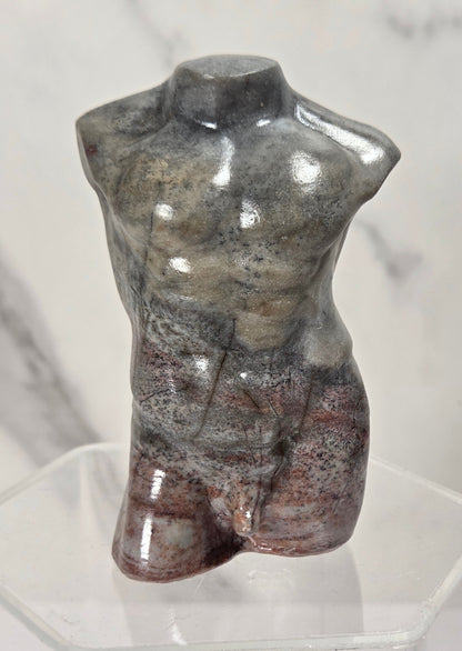 A polished stone torso sculpture with a mottled gray, brown, and reddish surface, displayed on a clear acrylic stand against a light marble-patterned background.