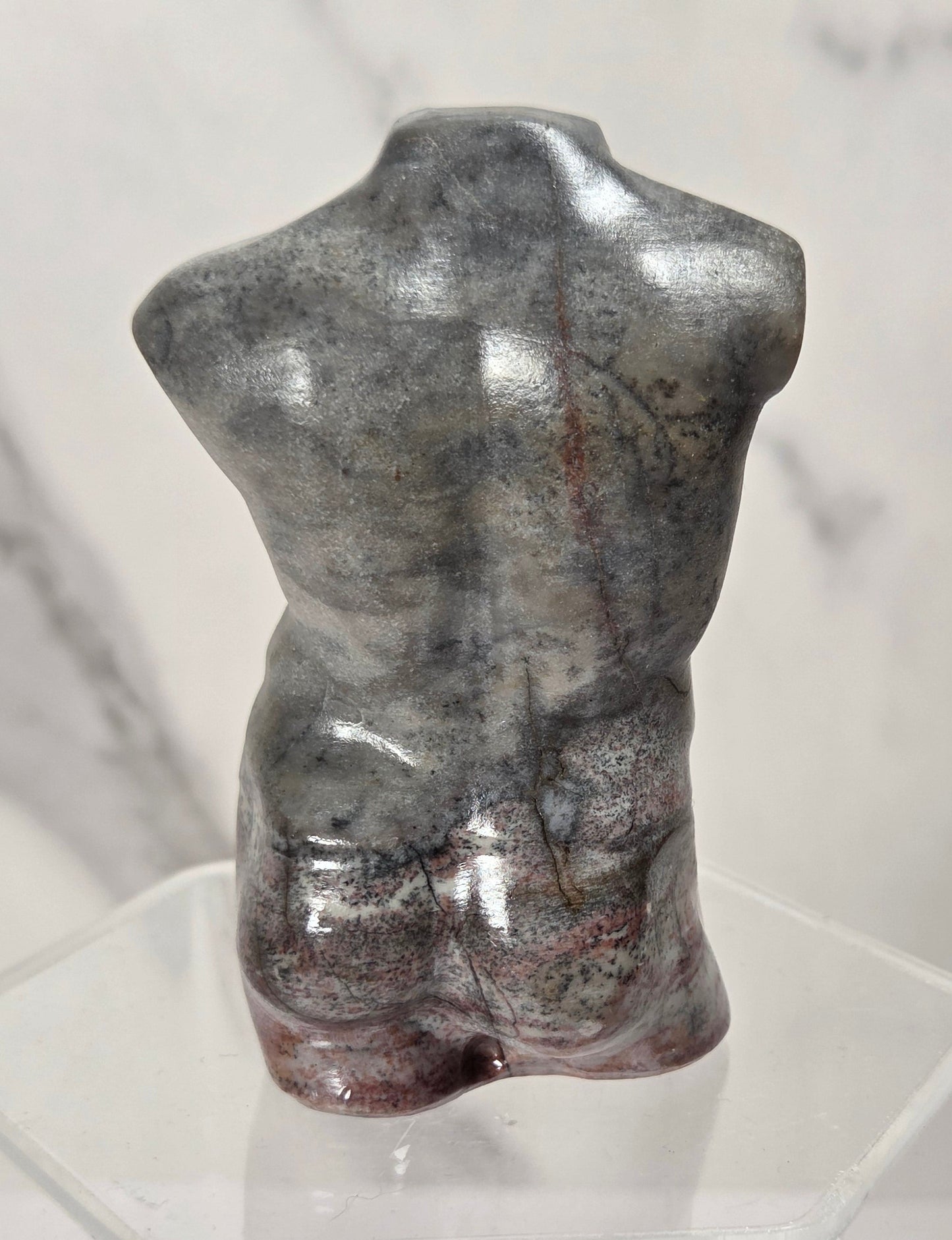 A small glossy torso sculpture in metallic gray with subtle reddish-brown marbling, displayed on a clear acrylic stand against a light marble-patterned background.