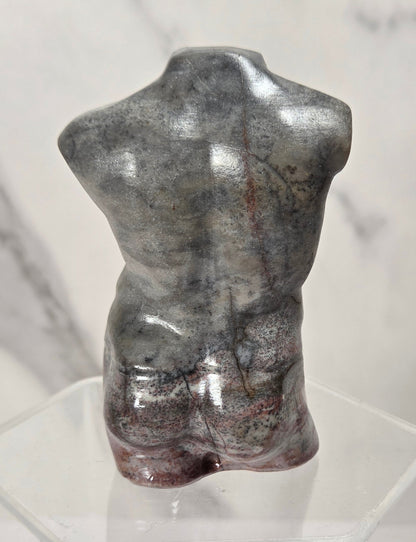 A small glossy torso sculpture in metallic gray with subtle reddish-brown marbling, displayed on a clear acrylic stand against a light marble-patterned background.