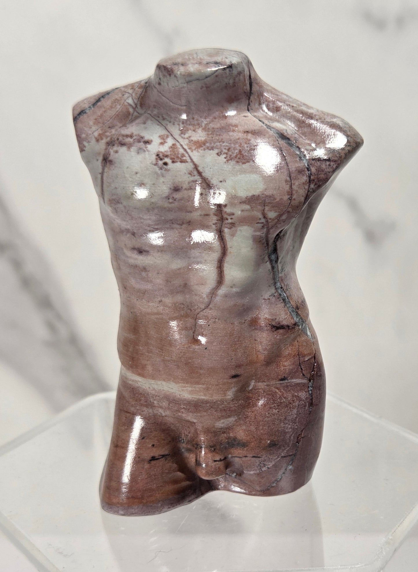 A glossy, sculptural torso mannequin made from polished brown and gray stone, displayed on a clear acrylic stand against a light marble-patterned background.