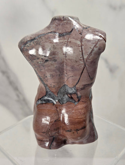 A polished reddish-brown stone torso sculpture with natural fissures, displayed on a clear acrylic pedestal against a light marble-patterned background.
