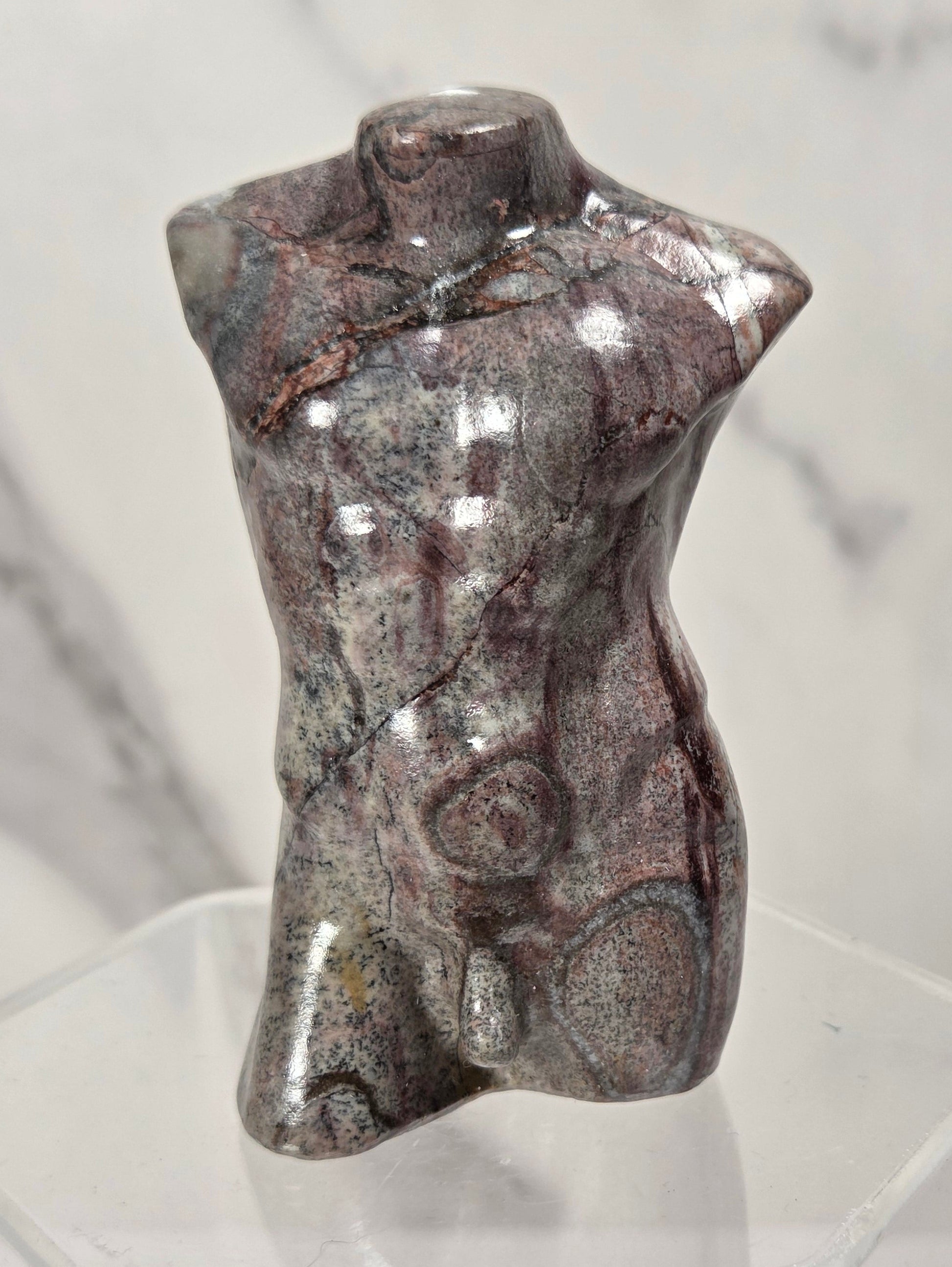A small polished stone torso sculpture with a glossy, metallic sheen, displayed on a clear acrylic stand against a light gray marble-patterned background.