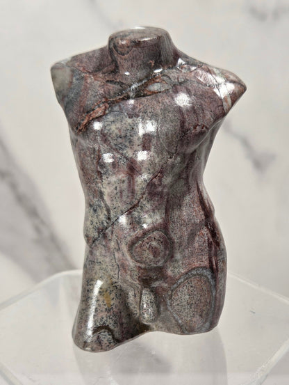 A small polished stone torso sculpture with a glossy, metallic sheen, displayed on a clear acrylic stand against a light gray marble-patterned background.