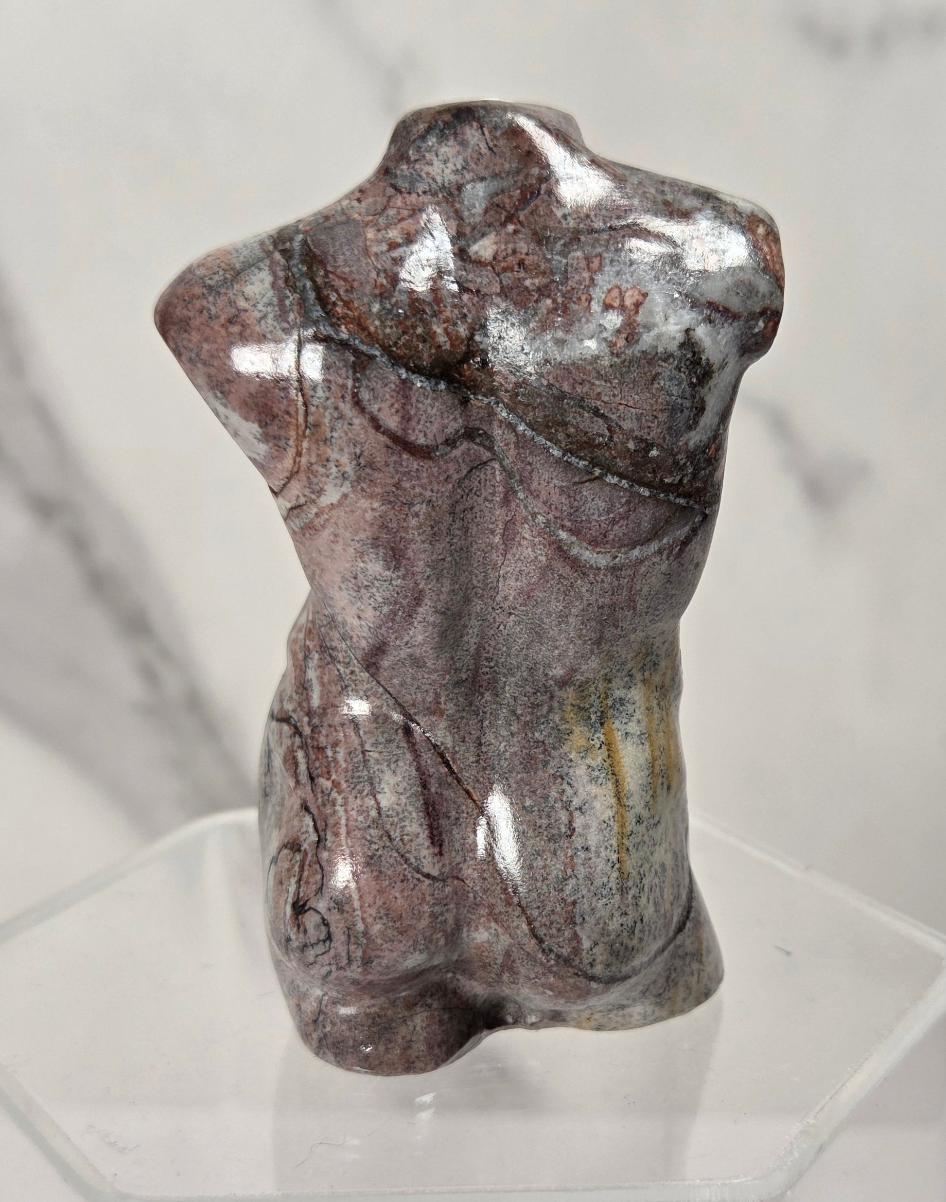 A polished decorative torso sculpture made from a marbled stone with brown, gray, and silver veining, displayed on a clear acrylic stand against a light gray marble-patterned background.