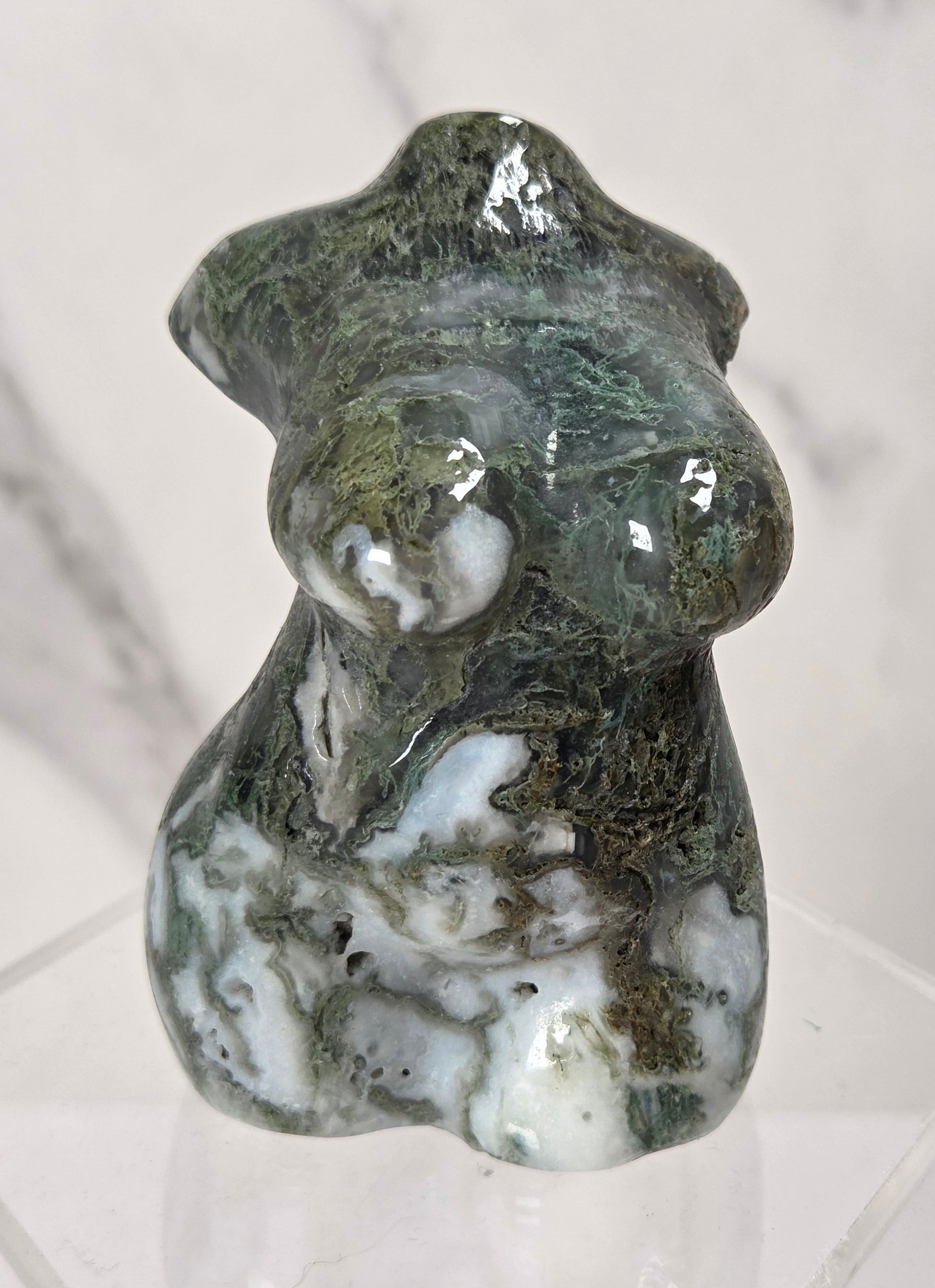 A polished natural stone figurine of a stylized female form, featuring green, white, and dark brown marbling, displayed on a clear acrylic stand against a light marble background.