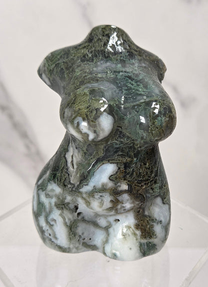 A polished natural stone figurine of a stylized female form, featuring green, white, and dark brown marbling, displayed on a clear acrylic stand against a light marble background.
