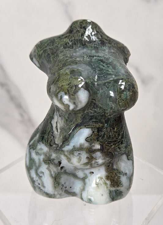 A polished natural stone figurine of a stylized female form, featuring green, white, and dark brown marbling, displayed on a clear acrylic stand against a light marble background.