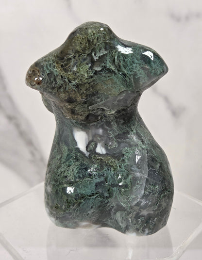 A small sculptural figurine of a seated figure with a glossy, mottled green and dark gray stone finish, displayed on a clear acrylic hexagonal pedestal against a light marble-patterned background.