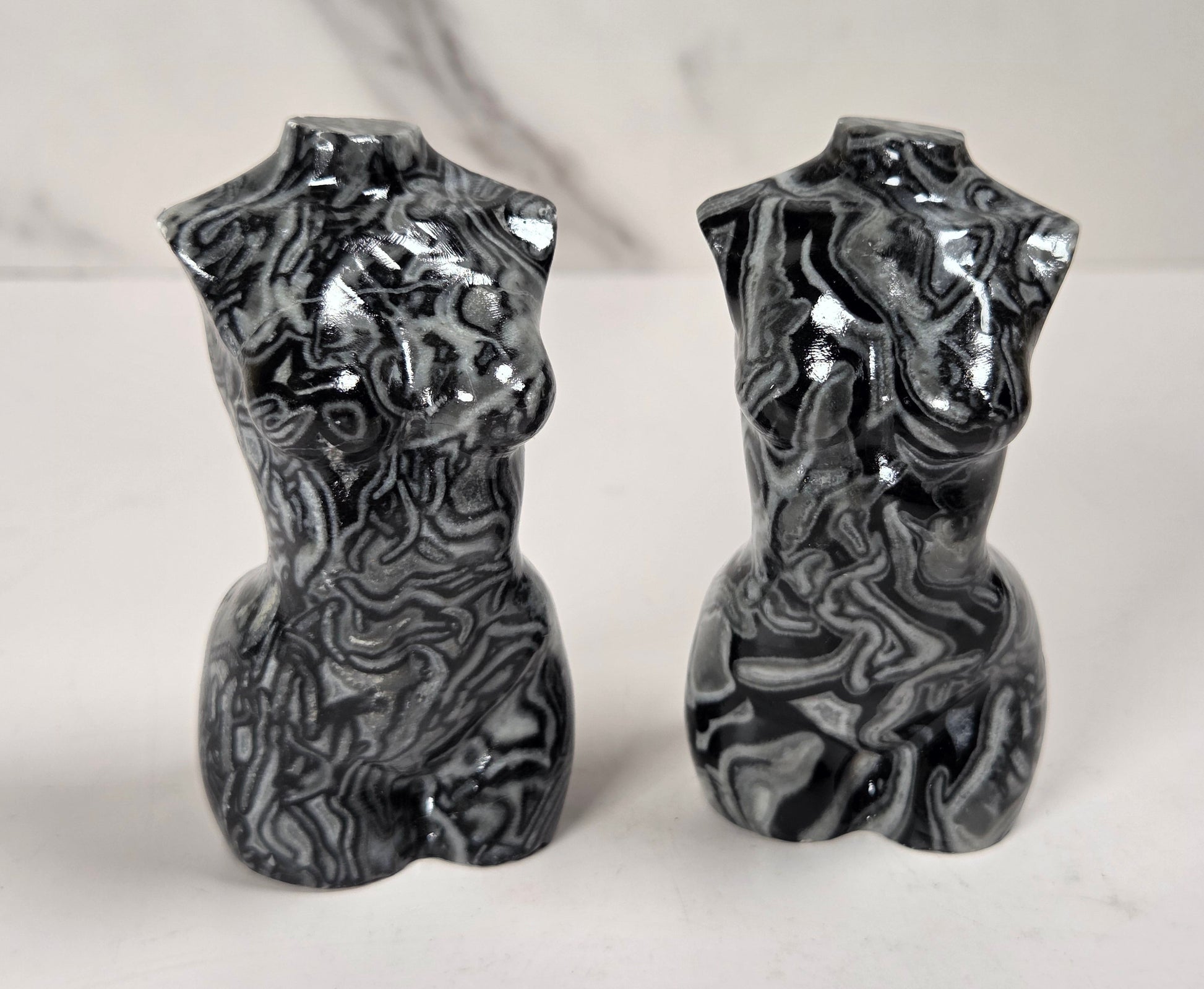 A pair of glossy black and white marbled torso-shaped ceramic vases or candle holders, standing side by side on a light surface against a white marble-patterned background.