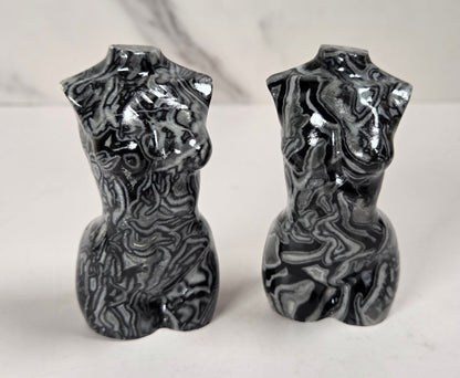 A pair of glossy black and white marbled torso-shaped ceramic vases or candle holders, standing side by side on a light surface against a white marble-patterned background.