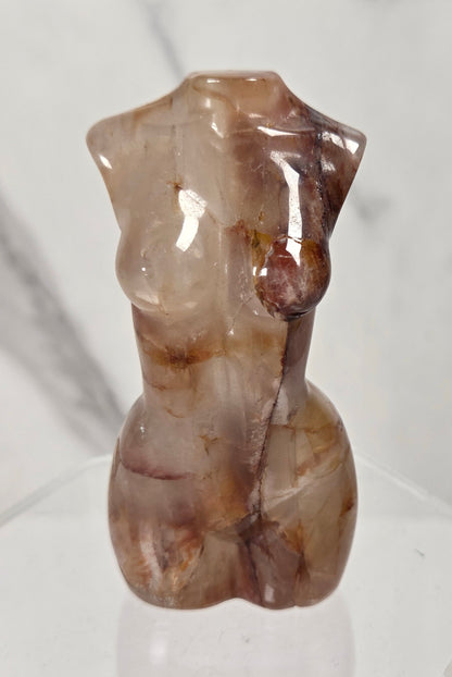 A small polished stone torso sculpture with a glossy finish, featuring natural brown and beige marbling, displayed on a clear acrylic hexagonal stand against a light gray marble-patterned background.