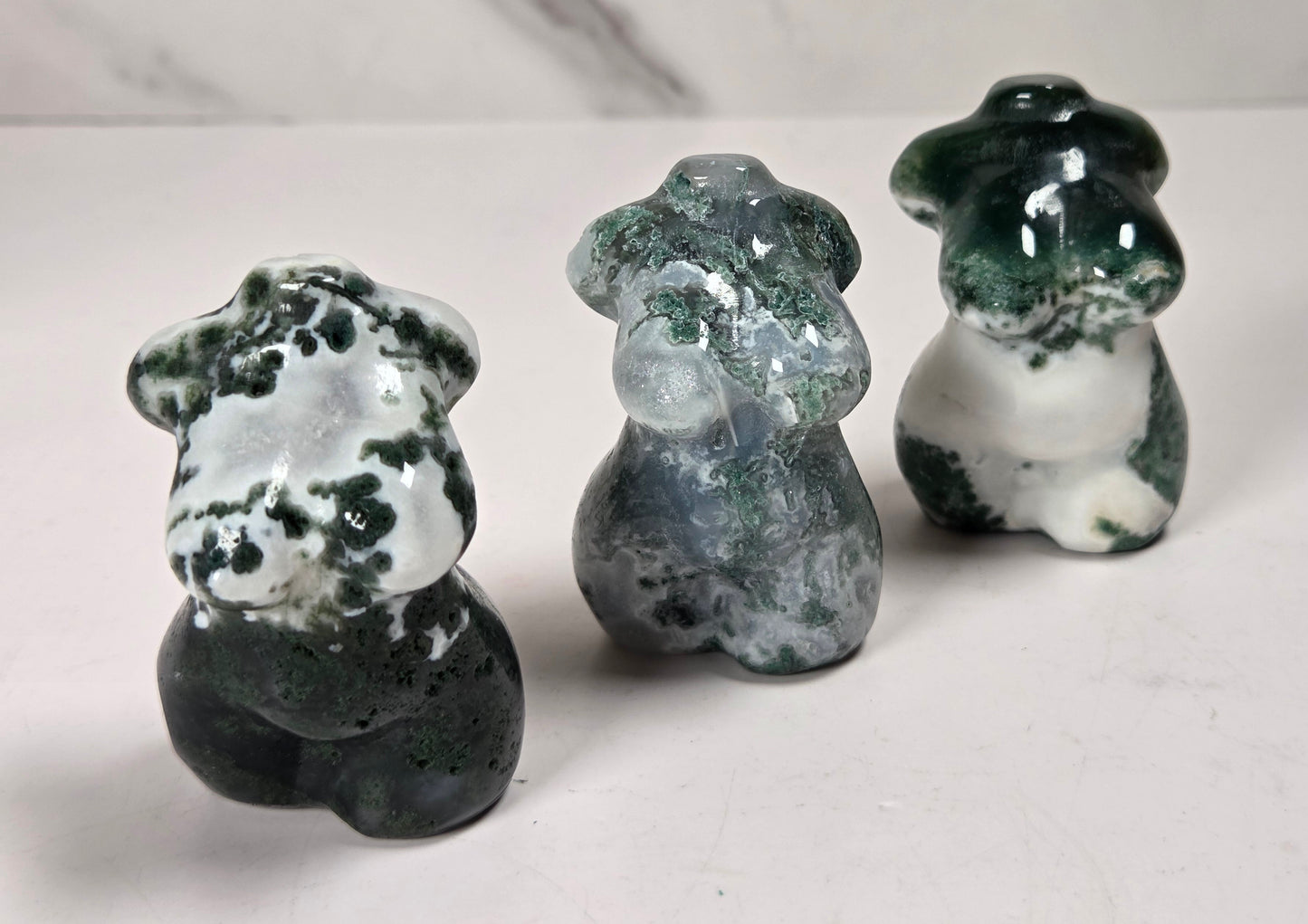 Three small carved stone figurines shaped like femail torsos, displayed on a light surface against a marble-look background. The figurines have marbled dark green, white, and gray stone patterns.