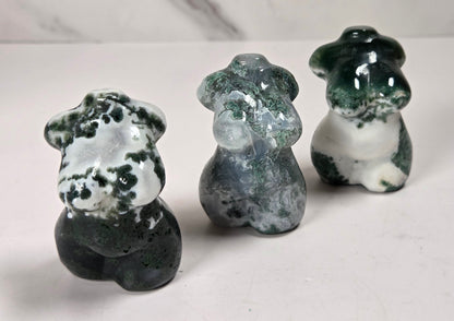 Three small carved stone figurines shaped like femail torsos, displayed on a light surface against a marble-look background. The figurines have marbled dark green, white, and gray stone patterns.