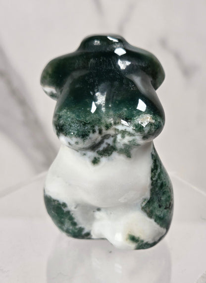A small polished stone figurine shaped like a female torso, featuring dark green and white marbled patterning, displayed on a clear acrylic stand against a light marble-patterned background.