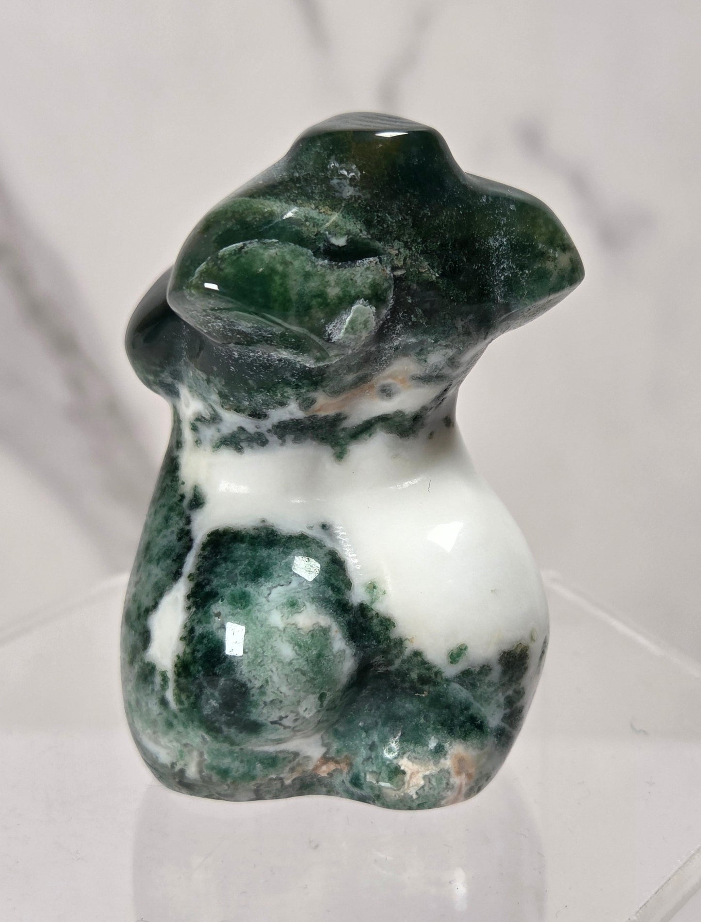 A small polished stone figurine shaped like a female torso, featuring dark green and white marbled patterning, displayed on a clear acrylic stand against a light marble-patterned background.