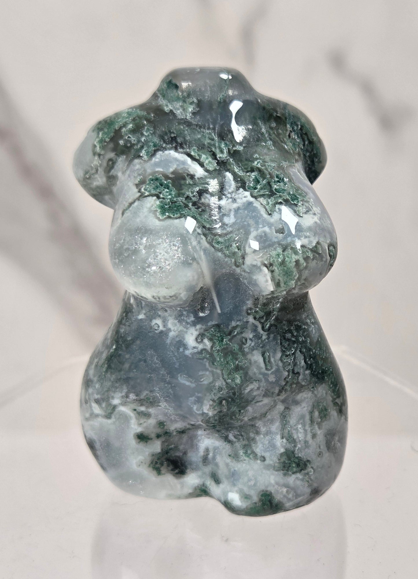 A small polished stone figurine shaped like a female torso, featuring dark green and white marbled patterning, displayed on a clear acrylic stand against a light marble-patterned background.